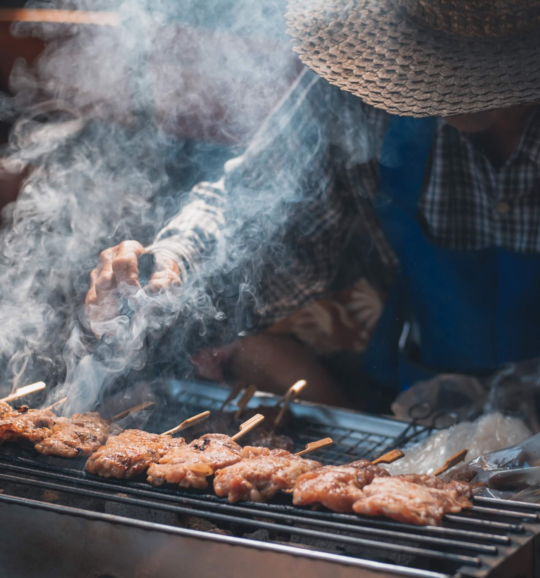 Image shows a man wearing a hat and grilling meat on sticks